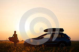 Dark silhouette of lonely woman relaxing near her car on grassy meadow enjoying view of colorful sunrise. Young female