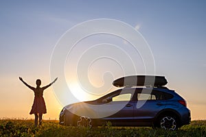 Dark silhouette of lonely woman relaxing near her car on grassy meadow enjoying view of colorful sunrise. Young female driver