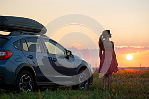 Dark silhouette of lonely woman relaxing near her car on grassy meadow enjoying view of colorful sunrise. Young female driver