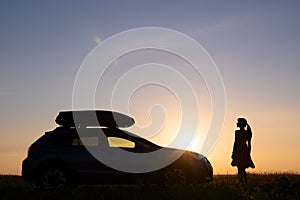 Dark silhouette of lonely woman relaxing near her car on grassy meadow enjoying view of colorful sunrise. Young female driver