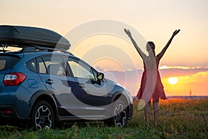 Dark silhouette of lonely woman relaxing near her car on grassy meadow enjoying view of colorful sunrise. Young female driver