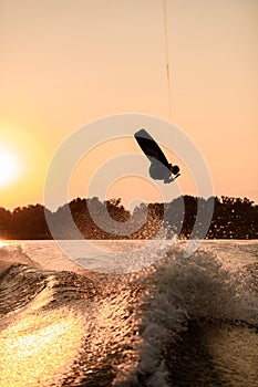 dark silhouette of man holding rope and making jump on wakeboard at sunset.