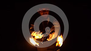 In the dark, a silhouette of a man is seen spinning flaming balls of fire on chains among the raindrops. Exciting fire