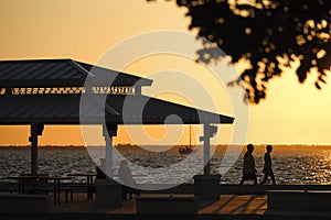 Dark silhouette of people resting under alcove roof on sea shore in public park at sunset