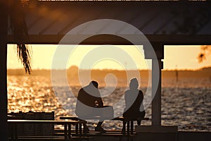 Dark silhouette of people resting under alcove roof on sea shore in public park at sunset