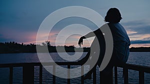 Dark silhouette of sad dreamy woman sitting on wooden rail of footbridge on lake at sunrise. Female person enjoying