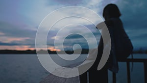 Dark silhouette of sad dreamy woman sitting on wooden rail of footbridge on lake at sunrise. Romantic vibe female person