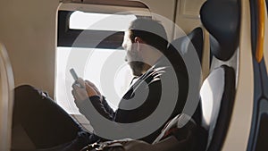 Dark silhouette of serious Caucasian man looking at his smartphone while traveling on the modern train window seat.