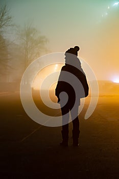 Dark silhouette of a woman in a hat with a backpack in the fog against the light of a city park. Vertical orientation.