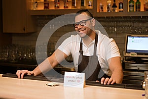 A dark-skinned bartender smiles at a visitor, there is a message on the bar counter prohibiting the sale of alcohol to