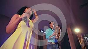 Dark-skinned black woman singer and thin saxophone player performing on stage.