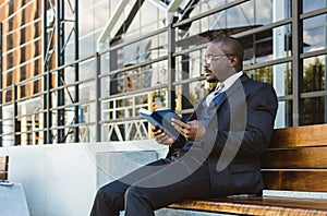 A dark-skinned businessman in a suit reads an outdoor notebook while sitting on a bench against the backdrop of city