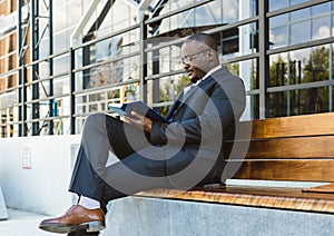 A dark-skinned businessman in a suit reads an outdoor notebook while sitting on a bench against the backdrop of city