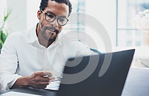 Dark skinned coworker wearing glasses and using laptop in modern office.African american man in white shirt working on