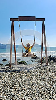 Dark-skinned Latino adult man with sunglasses, hat and shorts enjoys his vacation on the beach on a swing having fun and relaxing