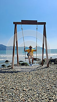 Dark-skinned Latino adult man with sunglasses, hat and shorts enjoys his vacation on the beach on a swing having fun and relaxing
