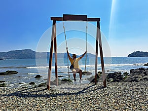 Dark-skinned Latino adult man with sunglasses, hat and shorts enjoys his vacation on the beach on a swing having fun and relaxing