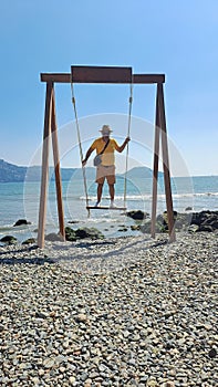 Dark-skinned Latino adult man with sunglasses, hat and shorts enjoys his vacation on the beach on a swing having fun and relaxing