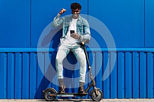 Dark-skinned man with electric scooter, using mobile phone , celebrating victory showing victory gesture