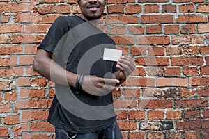 Dark-skinned man in T-shirt showing blank business card on bricked background.