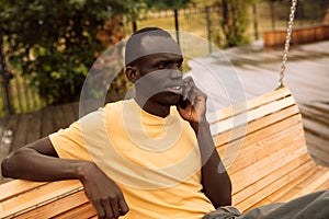 A dark-skinned man in a yellow T-shirt talks on the phone on a summer day in the park.