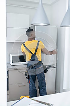 Dark-skinned service man in yellow tshirt repairing the fridge