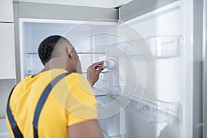 Dark-skinned service man in yellow tshirt repairing the fridge