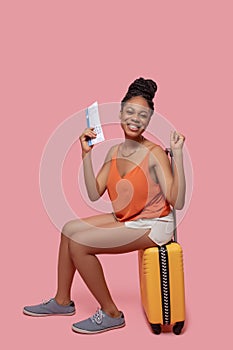 Dark-skinned woman holding a suitcase and a plane ticket and looking anticipated