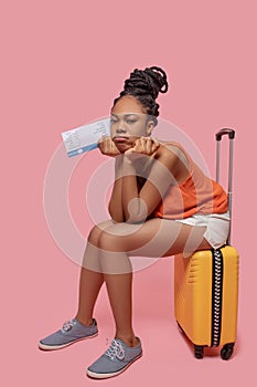 Dark-skinned woman holding a suitcase and a plane ticket and looking anticipated