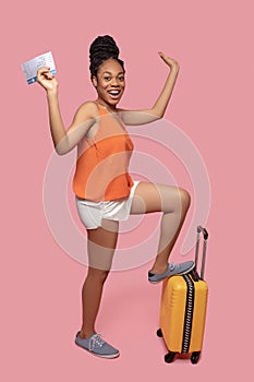 Dark-skinned woman holding a suitcase and a plane ticket and looking anticipated