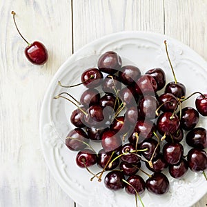 Dark sweet cherry in a white plate and on light wooden table close-up. Top view.