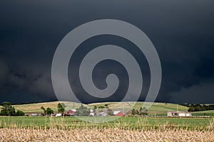 A dark wall cloud underneath a severe storm looms over a hill behind a farm.