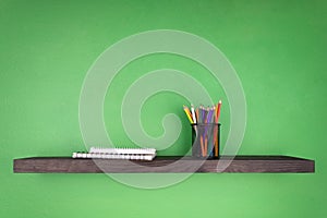 A dark wooden shelf against the background of a green wall on which is set a glass with pencils and notebooks with binding