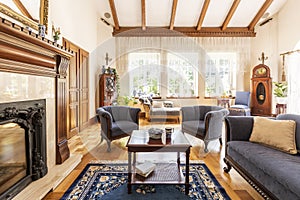 Dark wooden table on carpet between blue armchairs and sofa in luxury interior with fireplace. Real photo