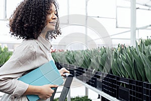 Darkskinned female farmer stands on stepladder among grown flowers