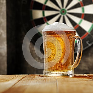 Dartboard with grey wall background. A big glass of cold beer on wooden table with empty space. Darts playing time.