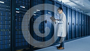 In Data Center: Male IT Specialist Wearing White Coat Stands Beside Server Rack, Uses Laptop Compu