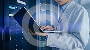 In Data Center: Male IT Specialist Wearing White Coat Stands Beside Server Rack, Uses Laptop Compu