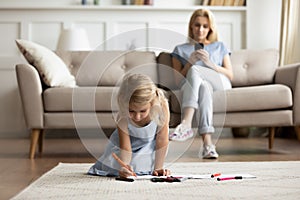 While daughter drawing seated on floor mom addicted with smartphone