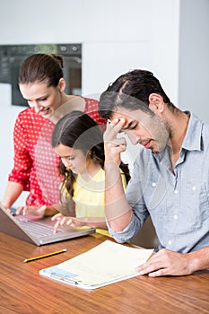 Daughter and mother working on laptop with tensed father sitting at desk