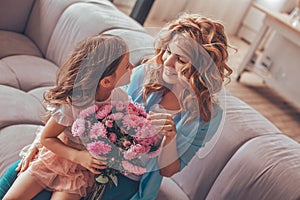 Daughter sitting on mothers laps with bouquet of flowers on the sofa and looking at each other