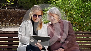 A daughter with a tablet and an elderly grandmother are sitting on a park bench.
