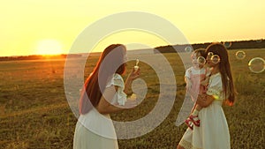 Daughters rejoice and smile, bubbles fly in park at sunset. Slow motion. Happy mother playing with children blowing soap