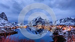 Dawn in icy lake and mountains of Norway, Reine next to lights of houses