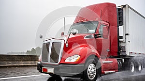 Day cab big rig red semi truck with roof spoiler transporting cargo in refrigerated semi trailer running on the wet rain highway