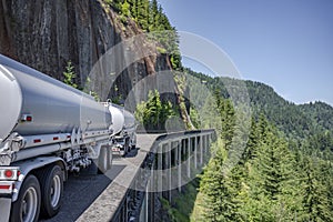 Day cab big rig semi truck transporting liquid cargo in two tank semi trailers running on the winding mountain road with rock wall