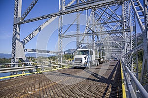 Day cab powerful big rig white semi truck with empty step down semi trailer driving on the silver truss bridge across Columbia