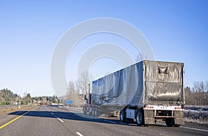 Day cab red big rig semi truck transporting commercial cargo in black covered semi trailer running on the straight wide highway