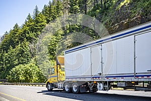 Day cab yellow big rig semi truck with roof spoiler driving with dry van semi trailer on the spectacular highway road with forest