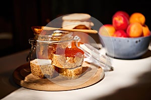 Daylight falls on part of the table with whole grain bread with apricot jam and ripe fruits in a bowl. Rustic still life